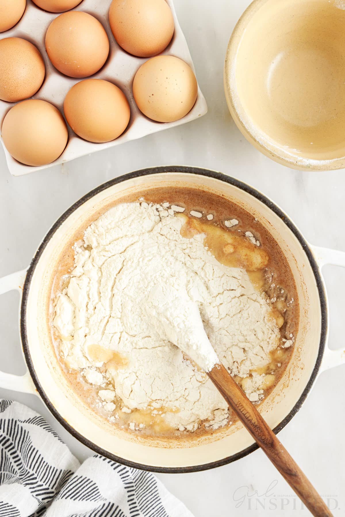 Flour added to melted butter mixture in Dutch oven with a wooden spoon inserted next to eggs and an empty dish to make Churro Recipe.