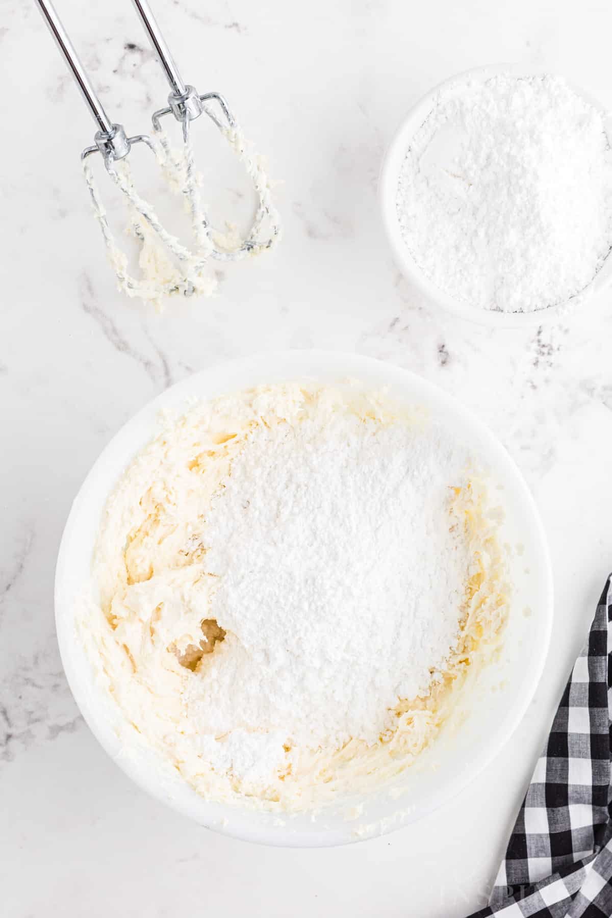 Bowl with cream cheese and butter mixture, powdered sugar added, hand mixer attachments coated in mixture.