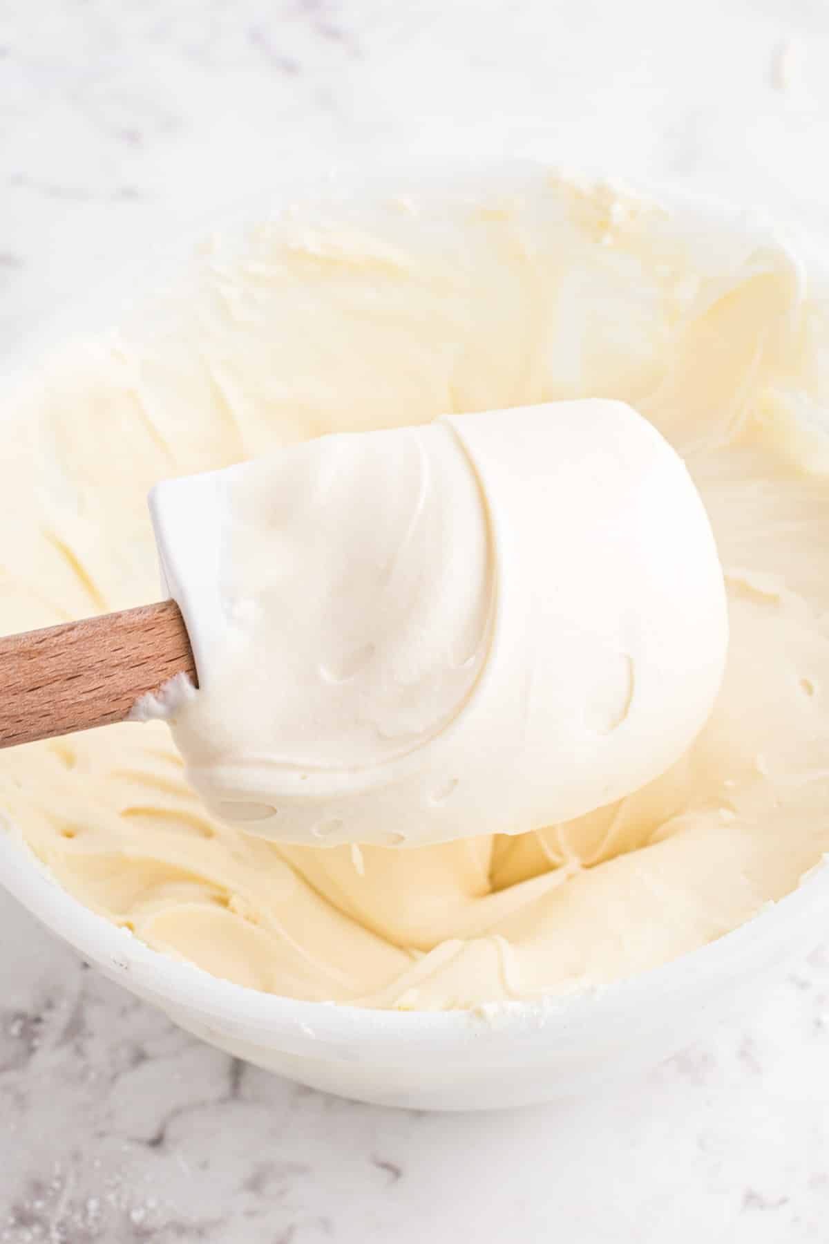 Plastic spatula held above a mixing bowl with cream cheese buttercream frosting.