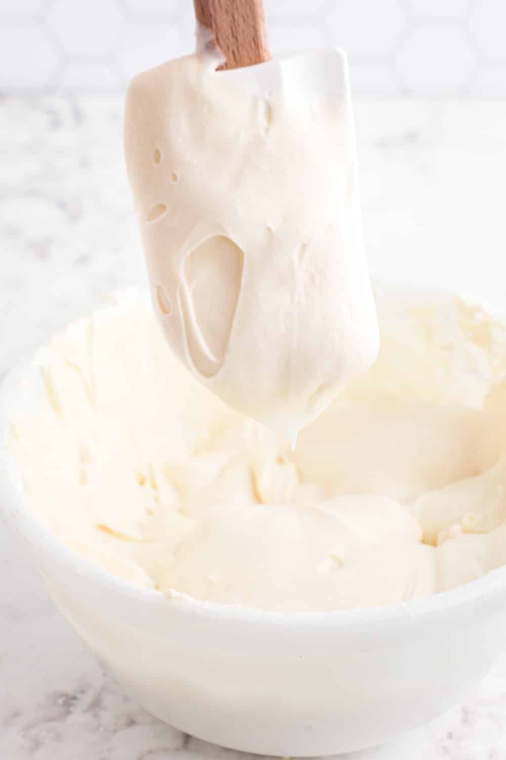 Cream cheese buttercream frosting in a mixing bowl, frosting on a plastic spatula held above the mixing bowl.