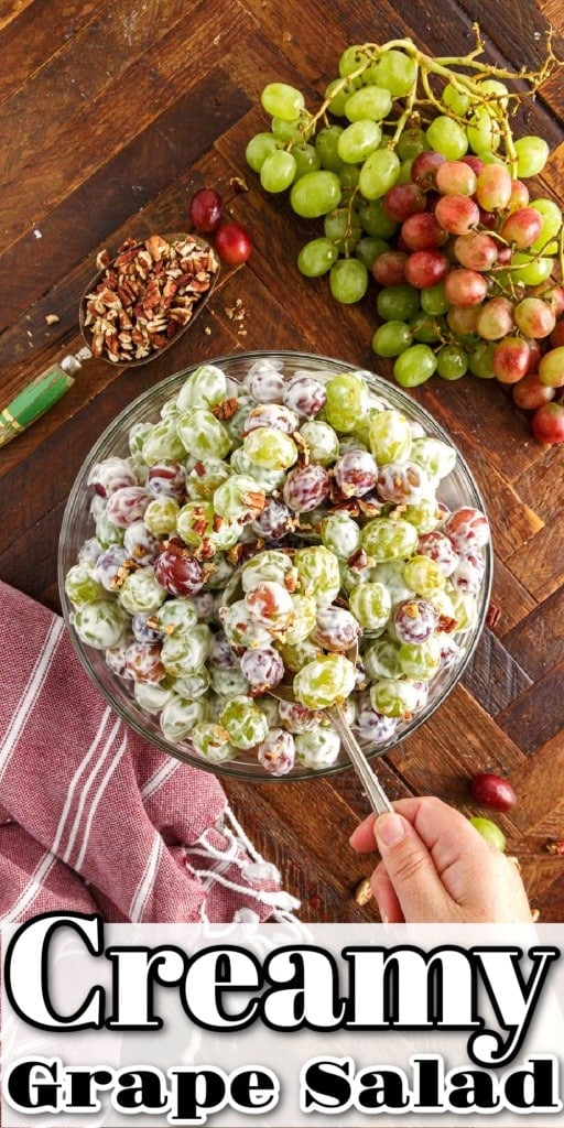 Overhead shot of creamy grape salad in a glass bowl on a wooden countertop with a spoon being used to scoop out some salad