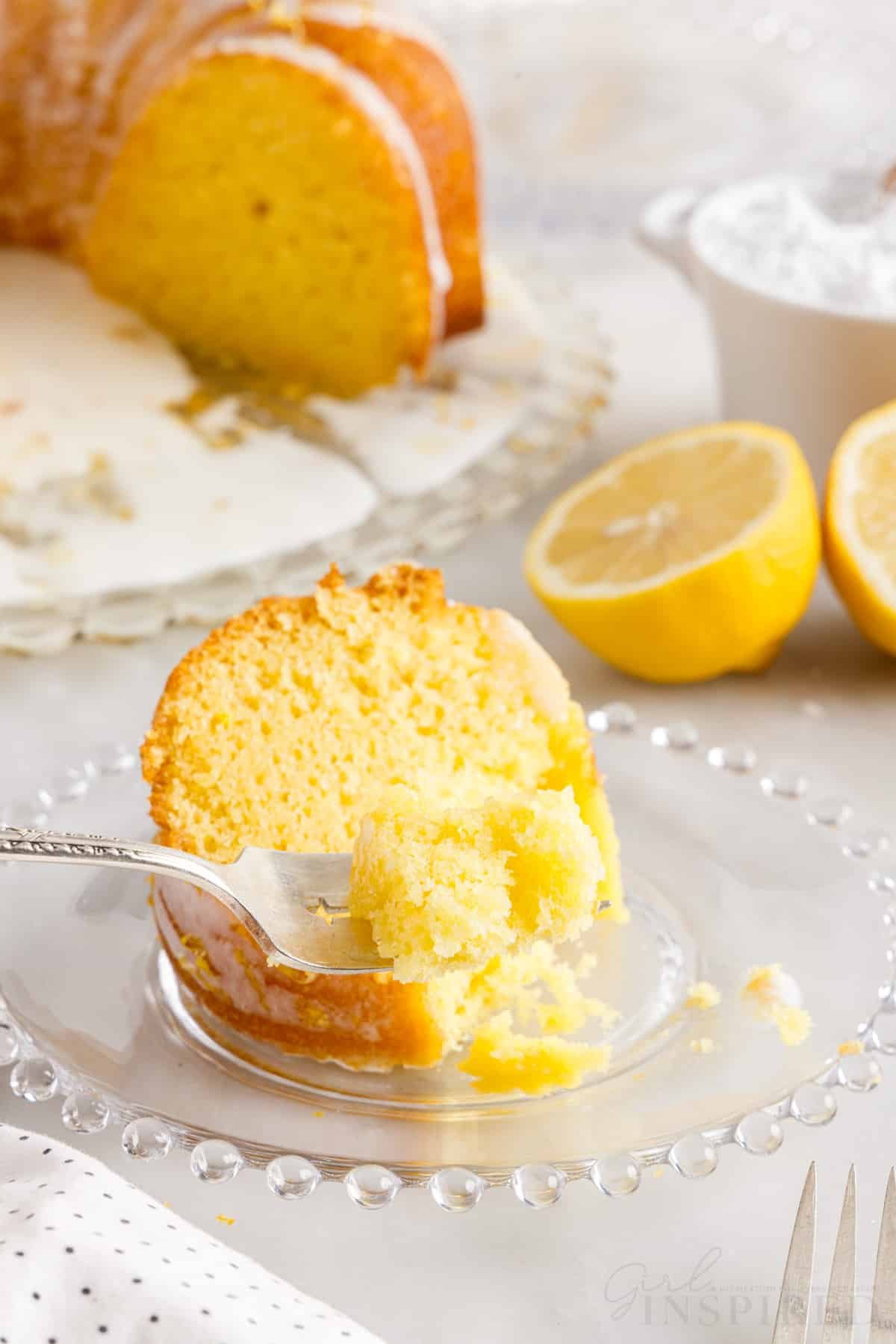 A small dish with a slice of Duncan Hines Lemon Cake on it, lemons and cake in the background.