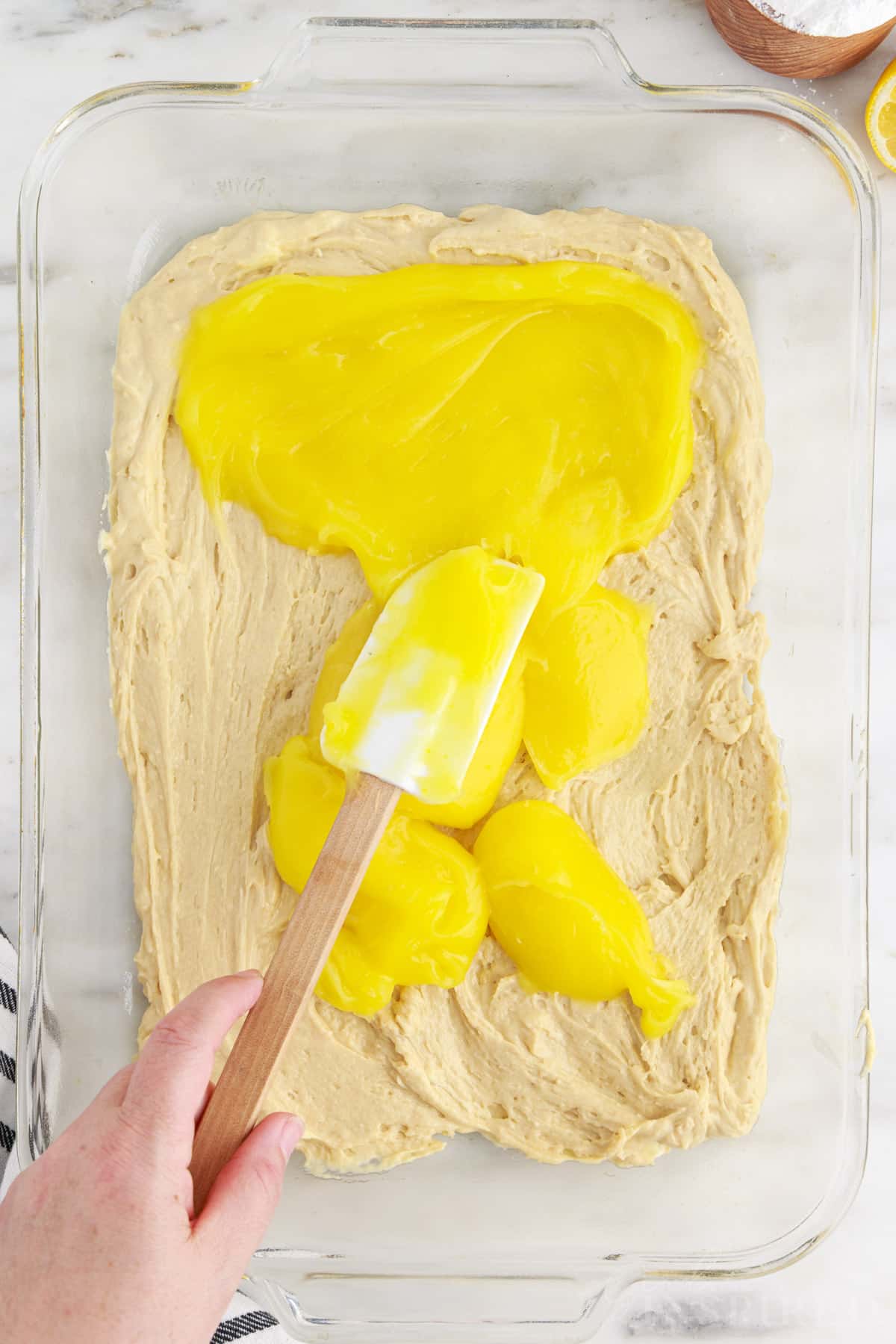 Hand holding a spatular that is spreading the lemon pie filling carefully over the first layer of lemon coffee cake batter. 
