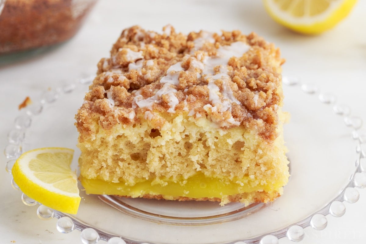 Close-up of a slice of lemon coffee cake on a glass serving plate and lemon slice garnish.