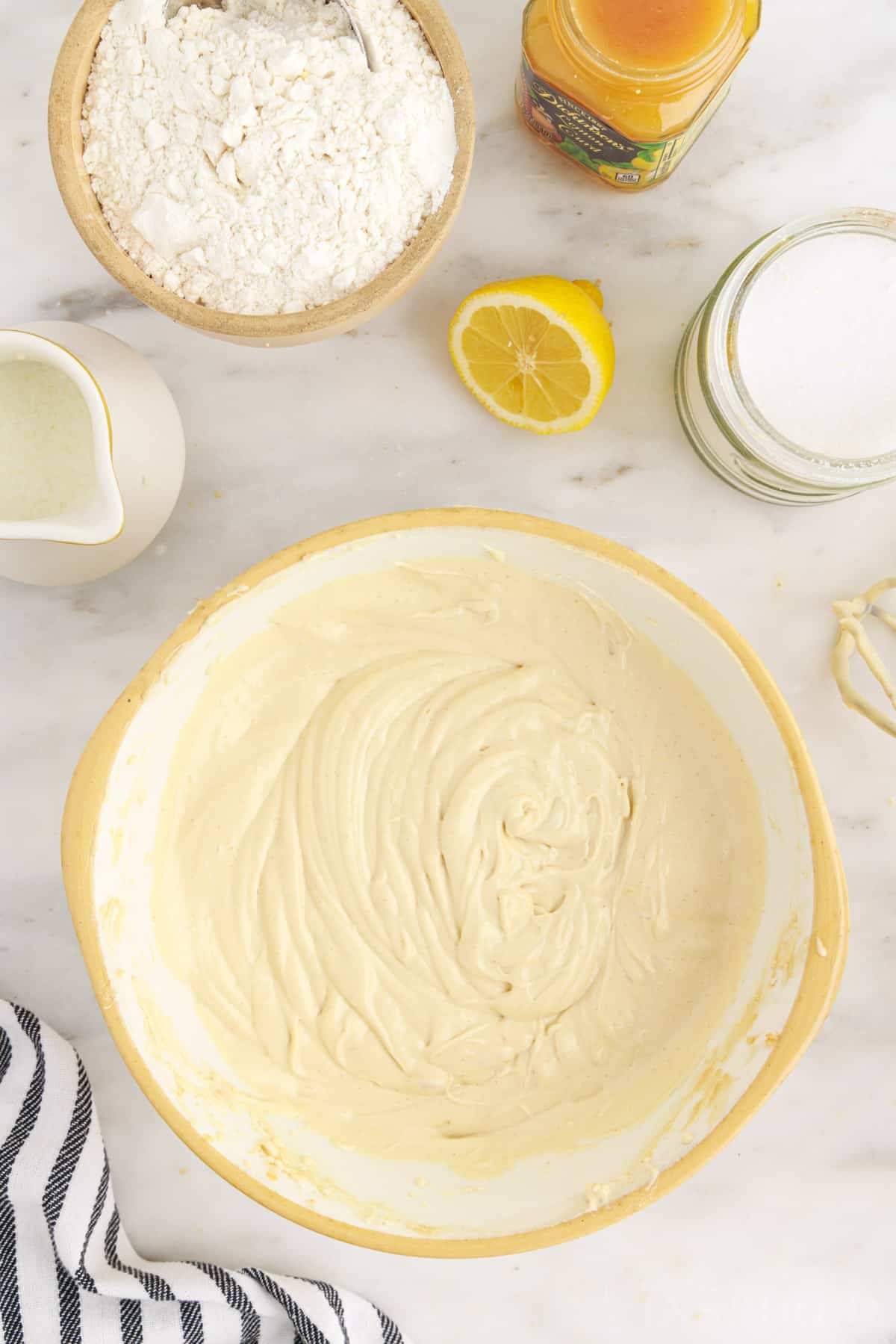 Bowl of combined cream cheese, butter, sugar, eggs, and vanilla extract, bowl of flour, bowl of baking soda and powder, bowl of cornstarch, jug of buttermilk, on a marble countertop.