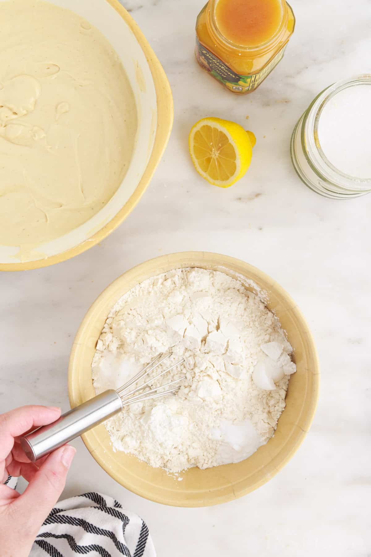 Bowl with flour, baking powder, baking soda, cornstarch, and salt, larger bowl of cake batter, jar of lemon pie filling, fresh sliced lemon, jar of buttermilk, on a marble countertop.