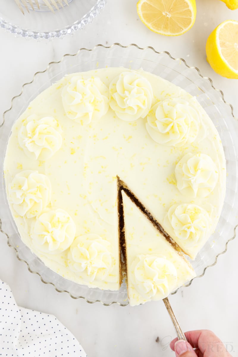 A slice of Lemon Layer Cake being removed from the Lemon Layer Cake next to lemons.