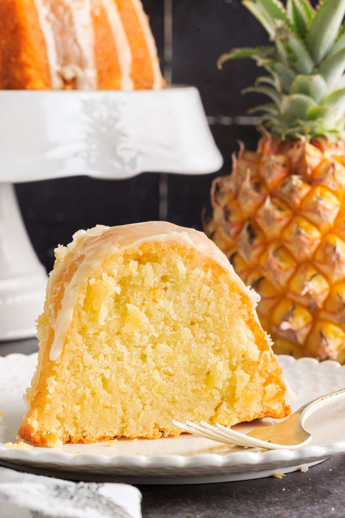 A slice of Pineapple Pound Cake on a small white dish with a fork a pineapple in the background.