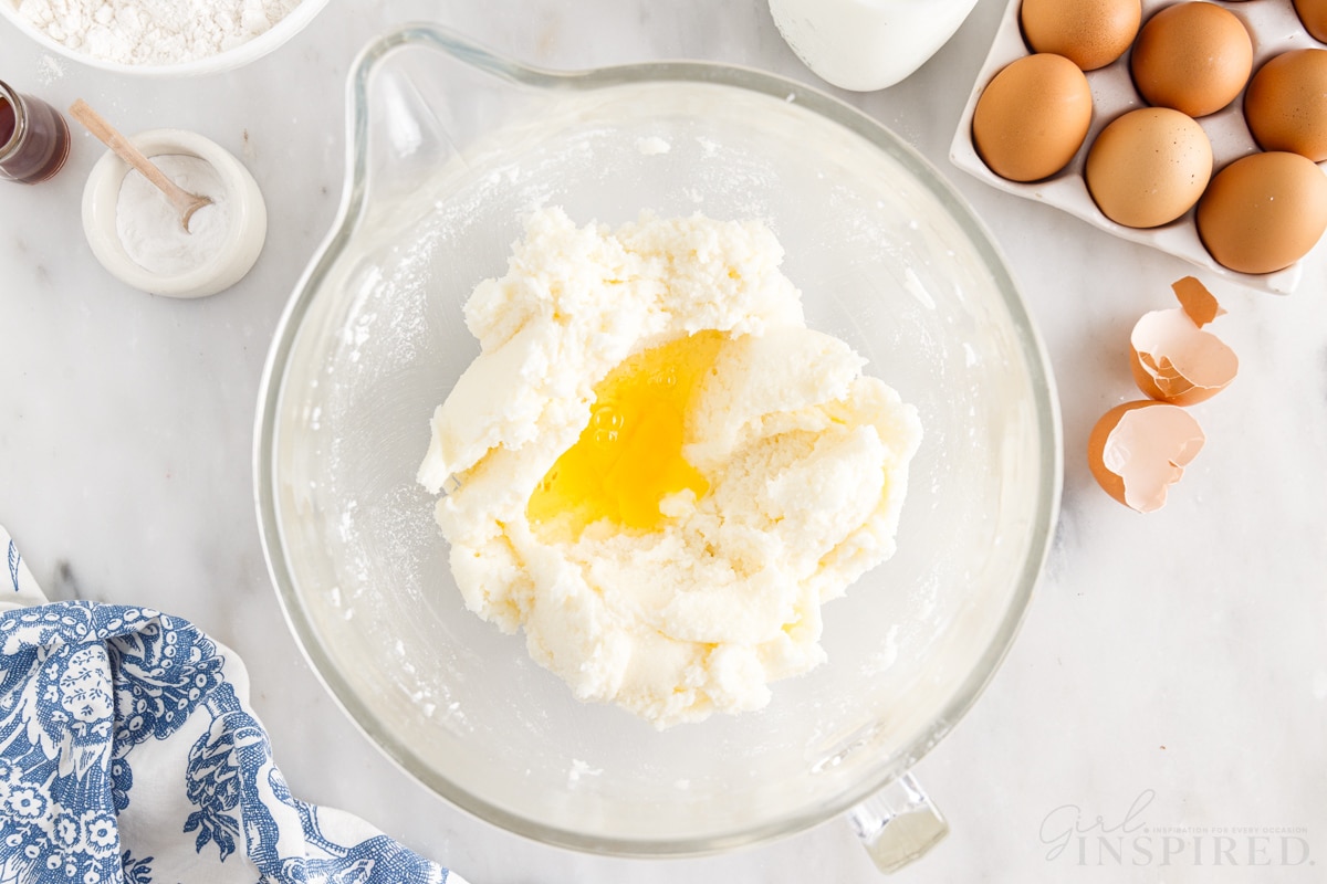 Eggs added to creamed butter mixture in a mixing bowl next to eggs and egg shells.