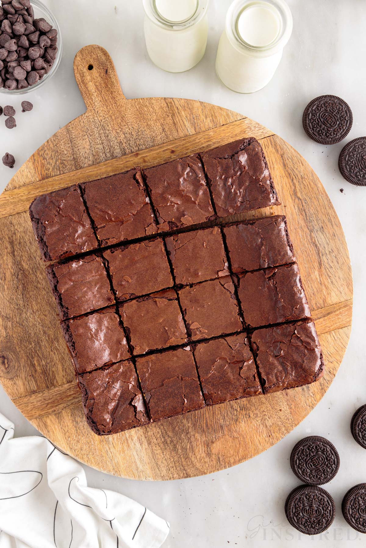 Slutty Brownies cut into squares on a round wooden cutting board next to chocolate chips, milk and Oreo´s.