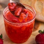 Close-up of strawberry freezer jam with metal spoon, bread and fresh strawberries in the background.