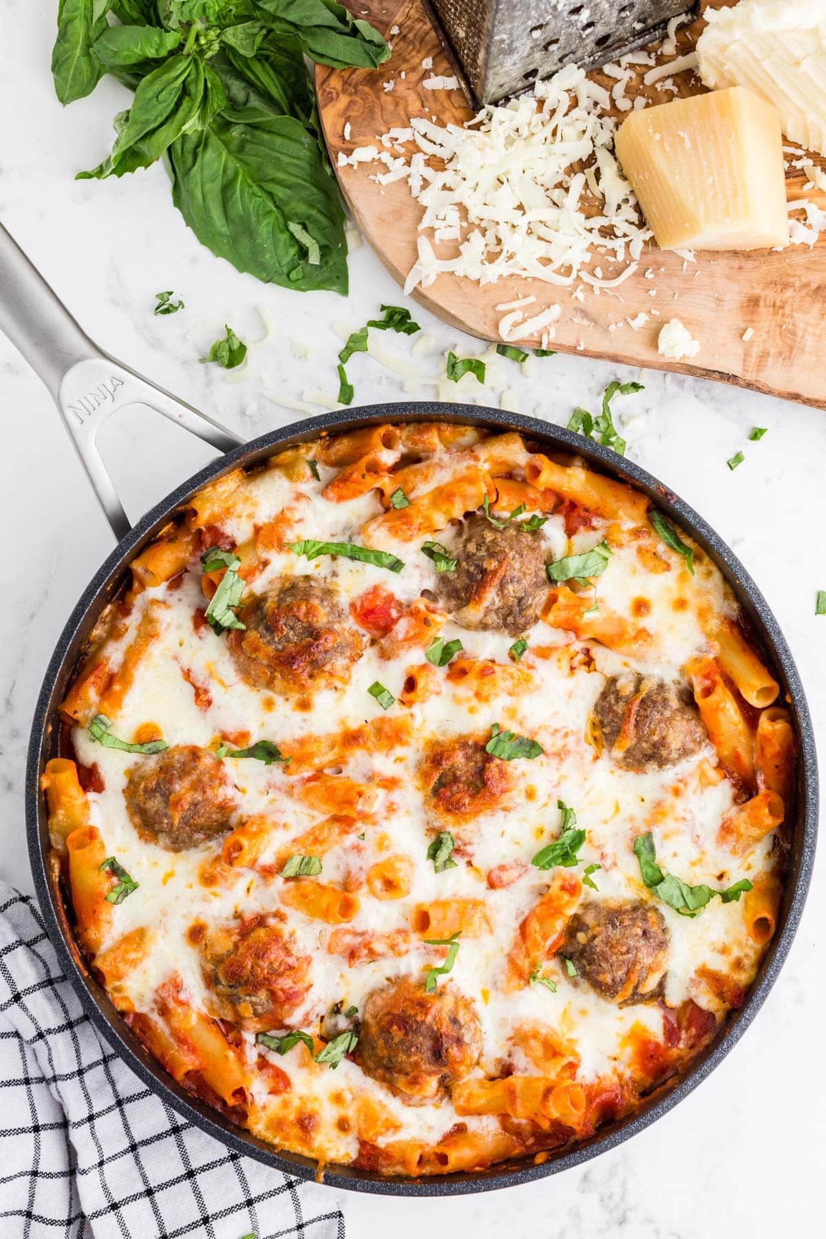 Baked Ziti with Meatballs in a large skillet on a marble countertop, wooden kitchen board with grated cheese and cheese blocks.