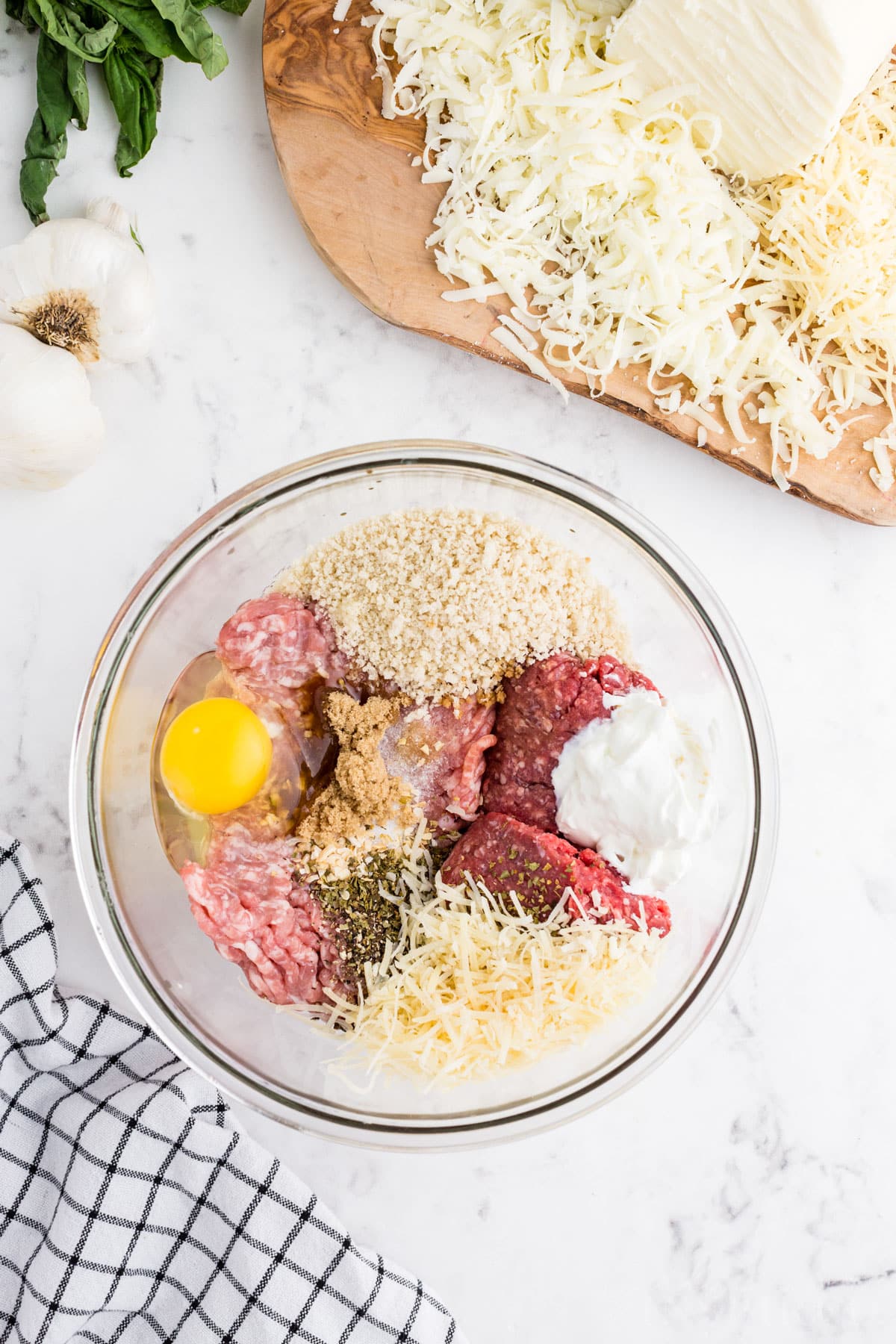 Medium mixing bowl with egg, ground meat, bread crumbs, seasoning, and parmesan cheese, wooden kitchen board with cheese, on a marble countertop.