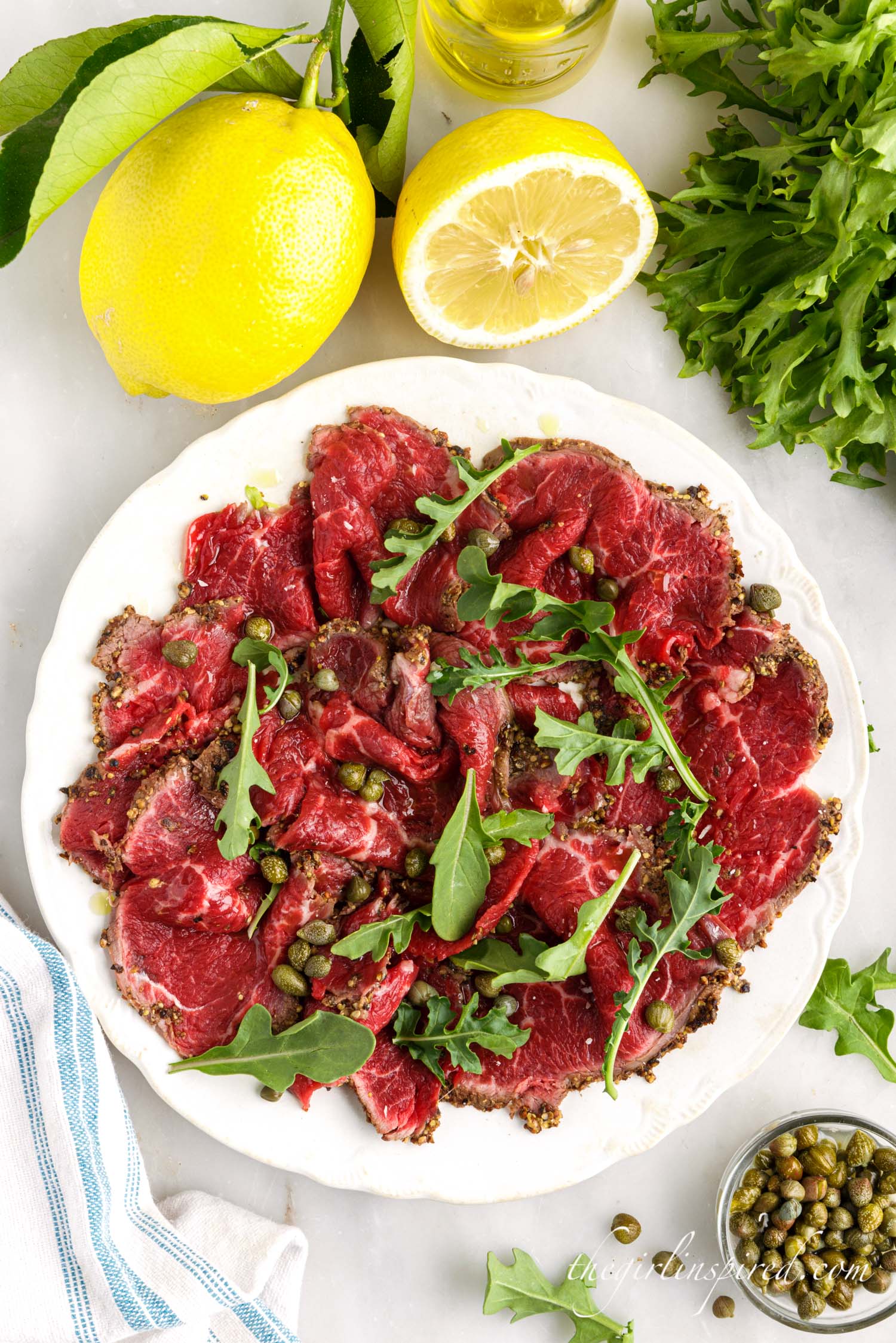 Beef Carpaccio on a plate next to capers, lemons, and arugula.