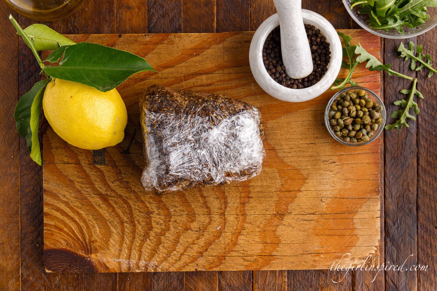 Browned tenderloin wrapped in plastic wrap on a cutting board next to a lemon and peppercorns.