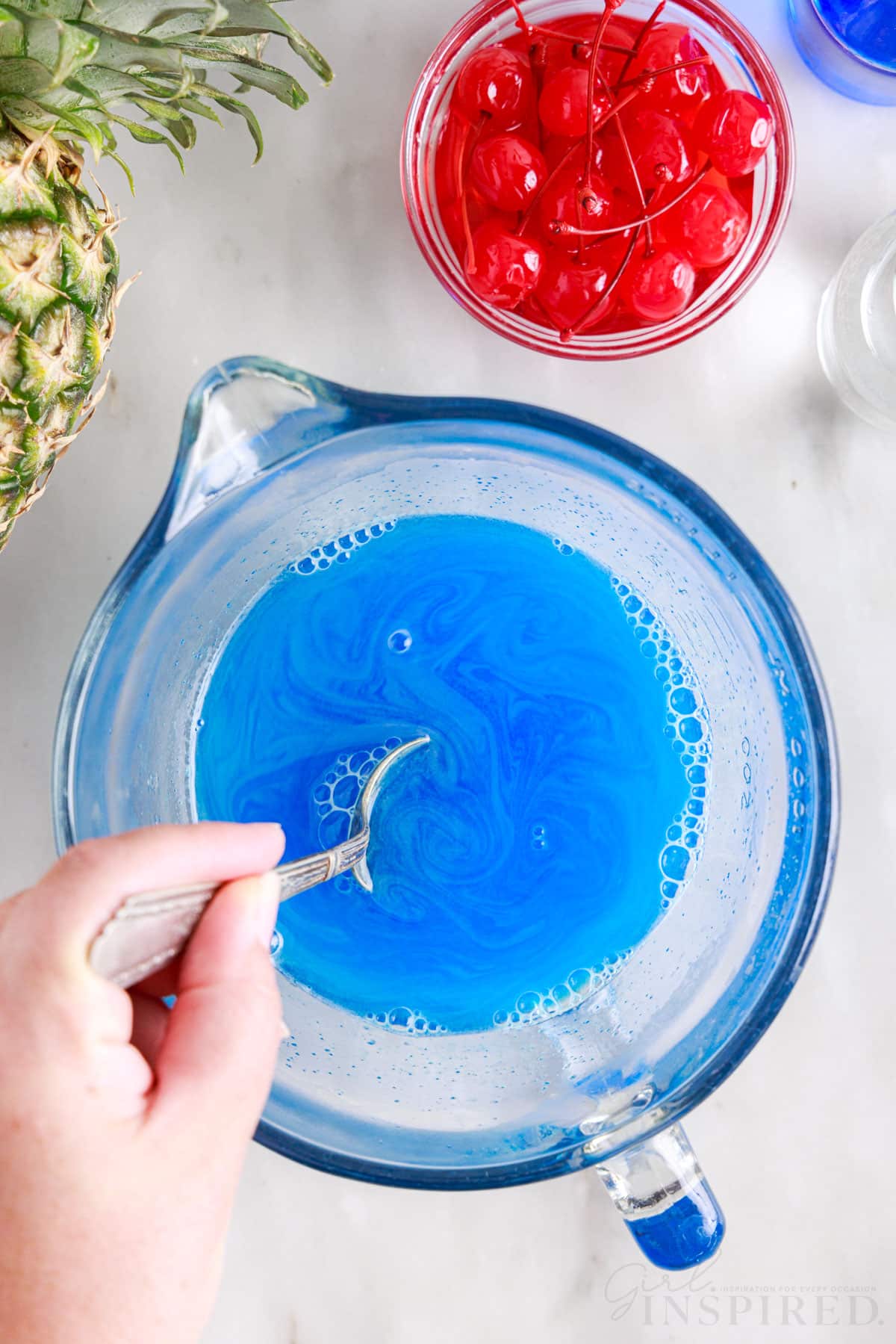 Boiling water stirred into blue jello with a spoon in a mixing bowl next to a bowl of cherries.