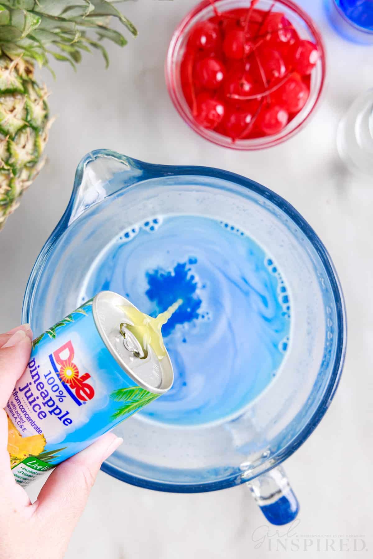 Pineapple juice added to blue jello mixture in a mixing bowl next to a pineapple and small dish of cherries.
