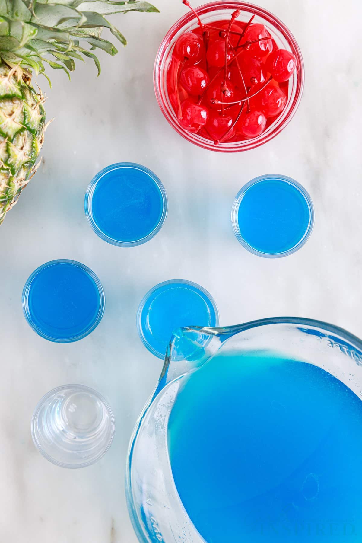 Blue Hawaiian Jello Shots being poured into shot glasses next to a dish of cherries.