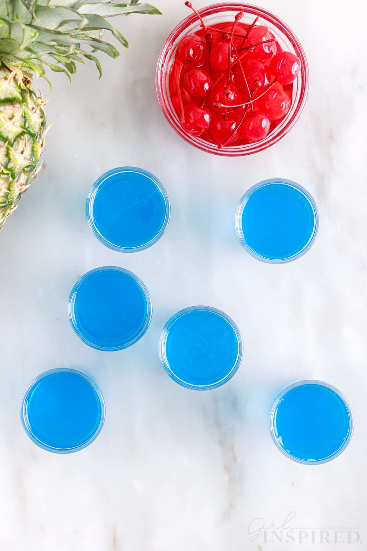 Six Blue Hawaiian Jello Shots next to cherries and a pineapple.