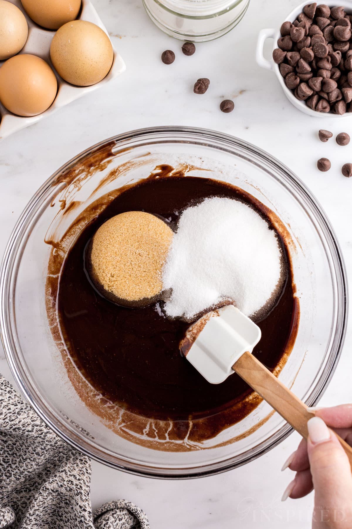 Mixing bowl with melted chocolate, butter, granulated sugar, and brown sugar with plastic spatula, brownie cupcake ingredients on a marble countertop.