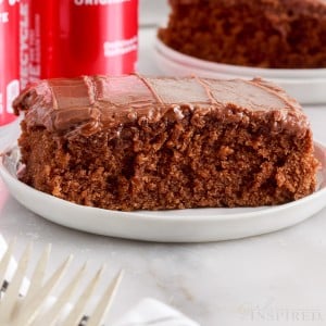 Front view of a slice of Coca Cola Cake on a small white dish, Cokes in the background.