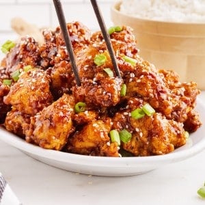 Close-up of General Tso Chicken with chopsticks, bowl of white rice in the background.