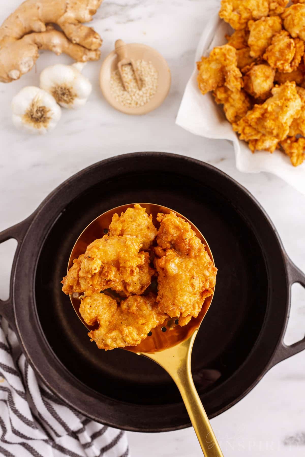 Slotted spoon with fried chicken pieces over a Dutch oven with hot oil, plate of freshly fried chicken pieces.