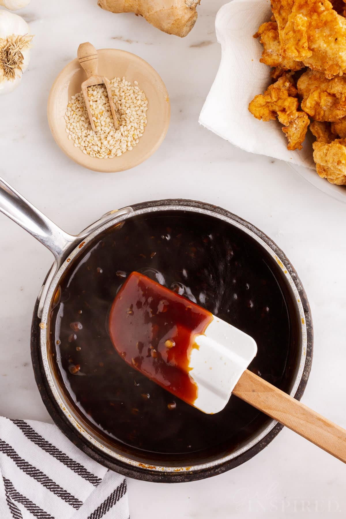 Saucepan with sticky sauce and plastic spatula, plate of fried chicken pieces.