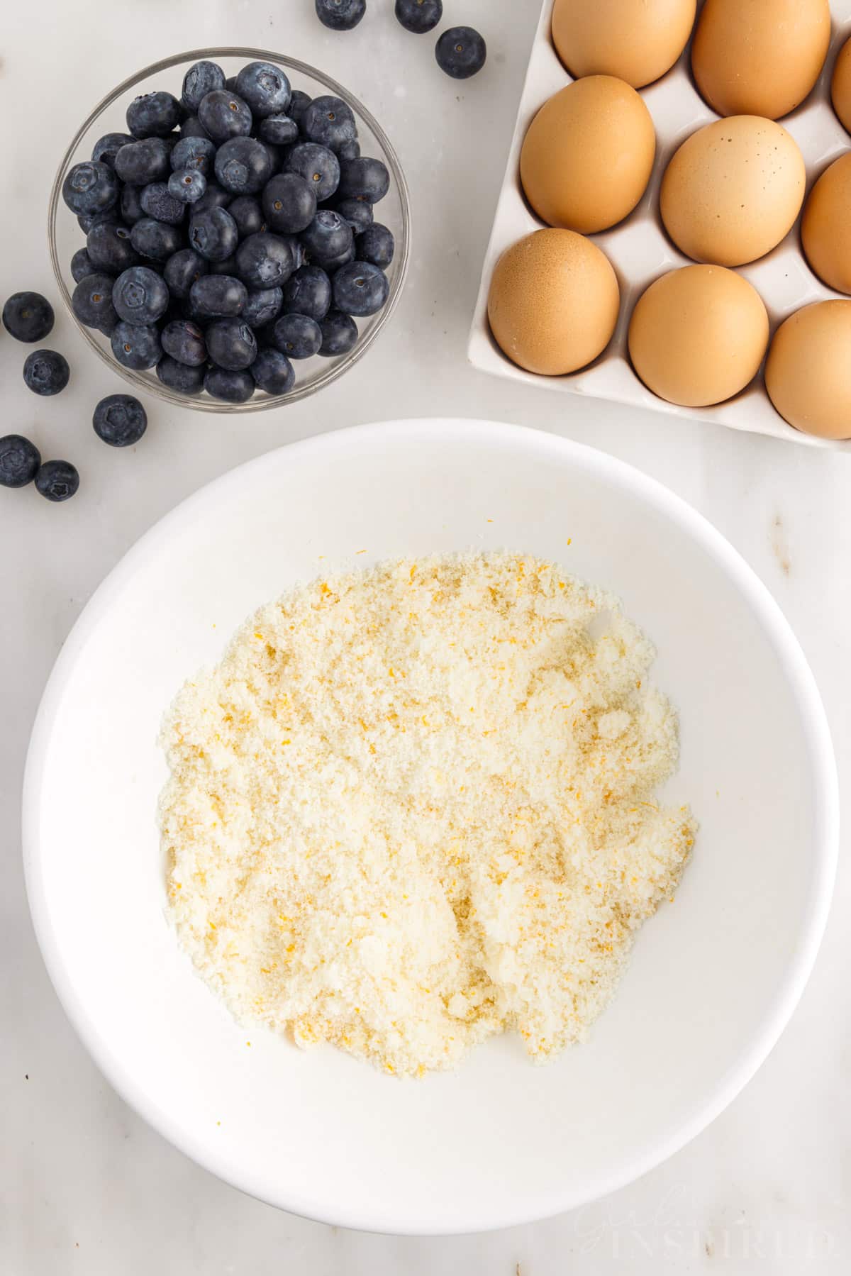 Lemon zest mixed into sugar in a mixing bowl next to eggs and blueberries.