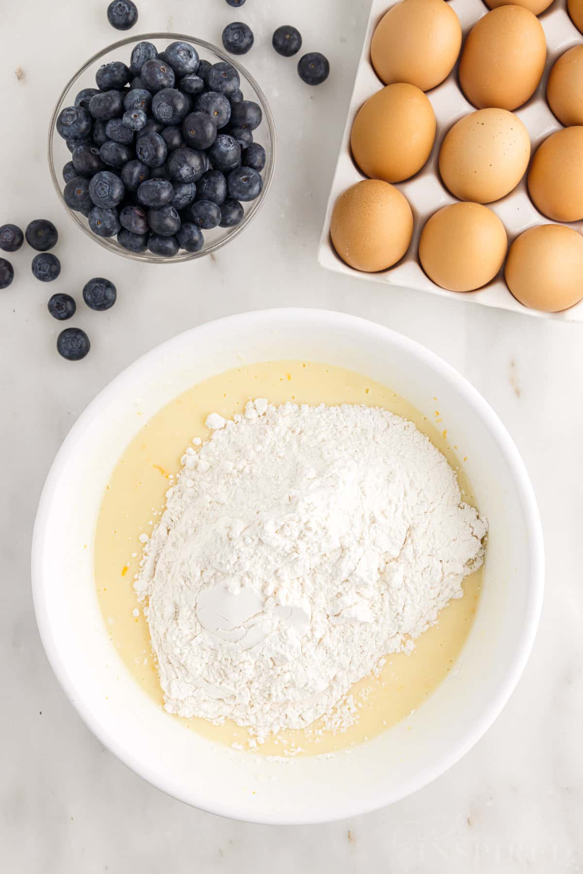 Dry ingredients are added to wet ingredients next to eggs and blueberries.