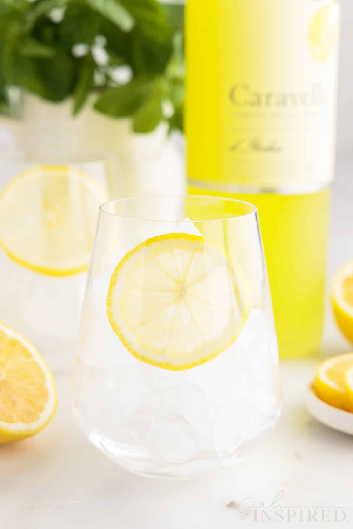 Two glasses of ice with lemon slices in them with a bottle of Limoncello in the background.