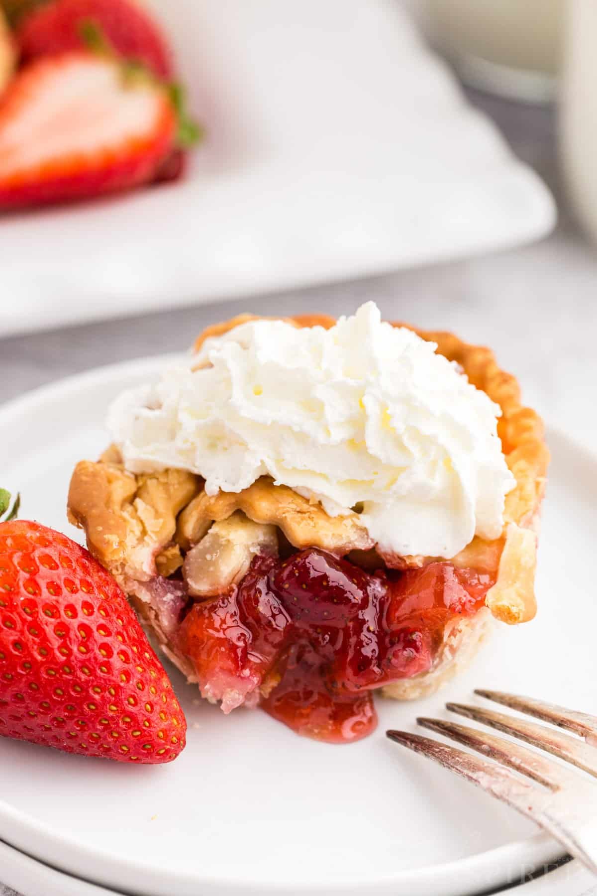 A strawberry rhubarb mini pie on a dessert plate with a bite removed and topped with whipped cream.