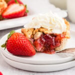 A strawberry rhubarb mini pie on a dessert plate with a bite removed and topped with whipped cream, a whole strawberry.