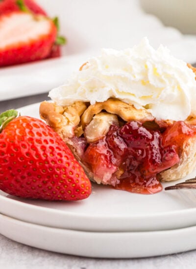 A strawberry rhubarb mini pie on a dessert plate with a bite removed and topped with whipped cream, a whole strawberry.