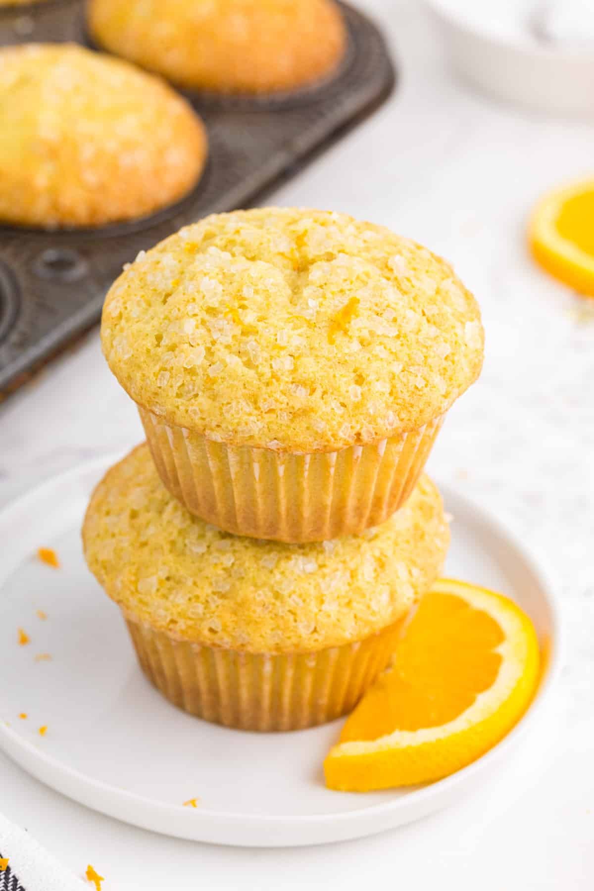 Two stacked Orange Muffins on a white serving plate, metal muffin pan with orange muffins.