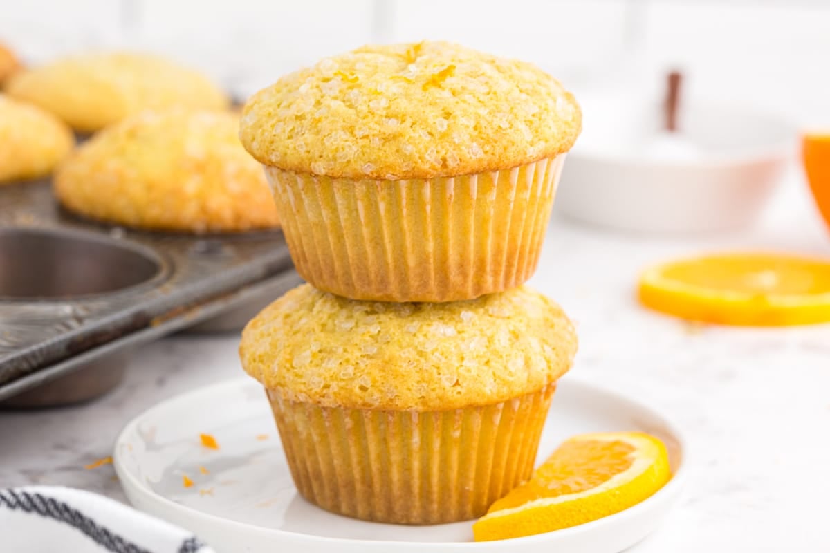 Two stacked orange muffins on a white serving plate, metal muffin tray with orange muffins in the background.