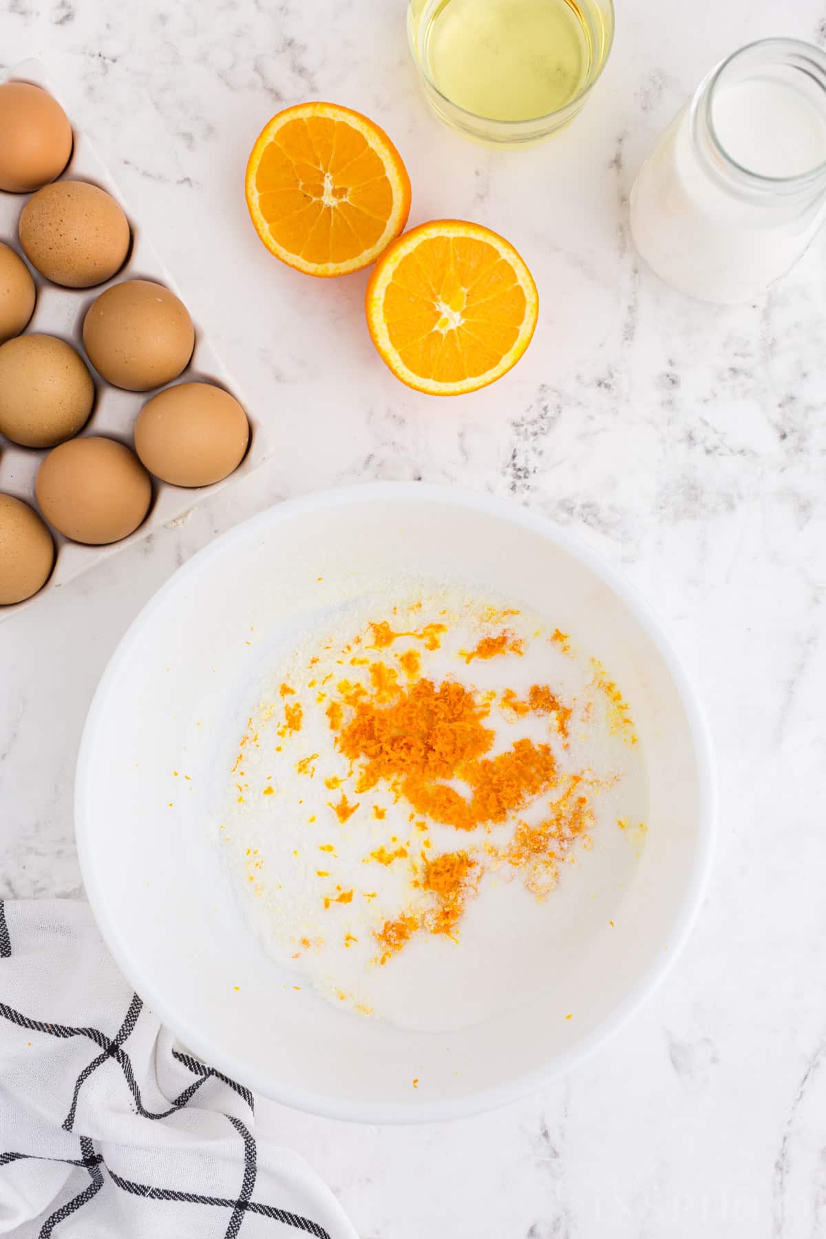 Large mixing bowl with granulated sugar and orange zest, fresh ingredients on a marble countertop.