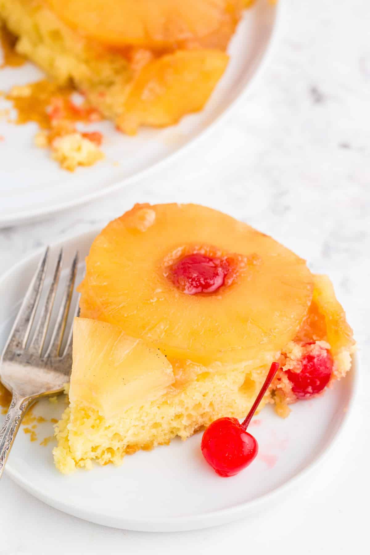 Slice of pineapple upside down cake on a serving plate with a metal fork, remaining pineapple upside down cake in the background.