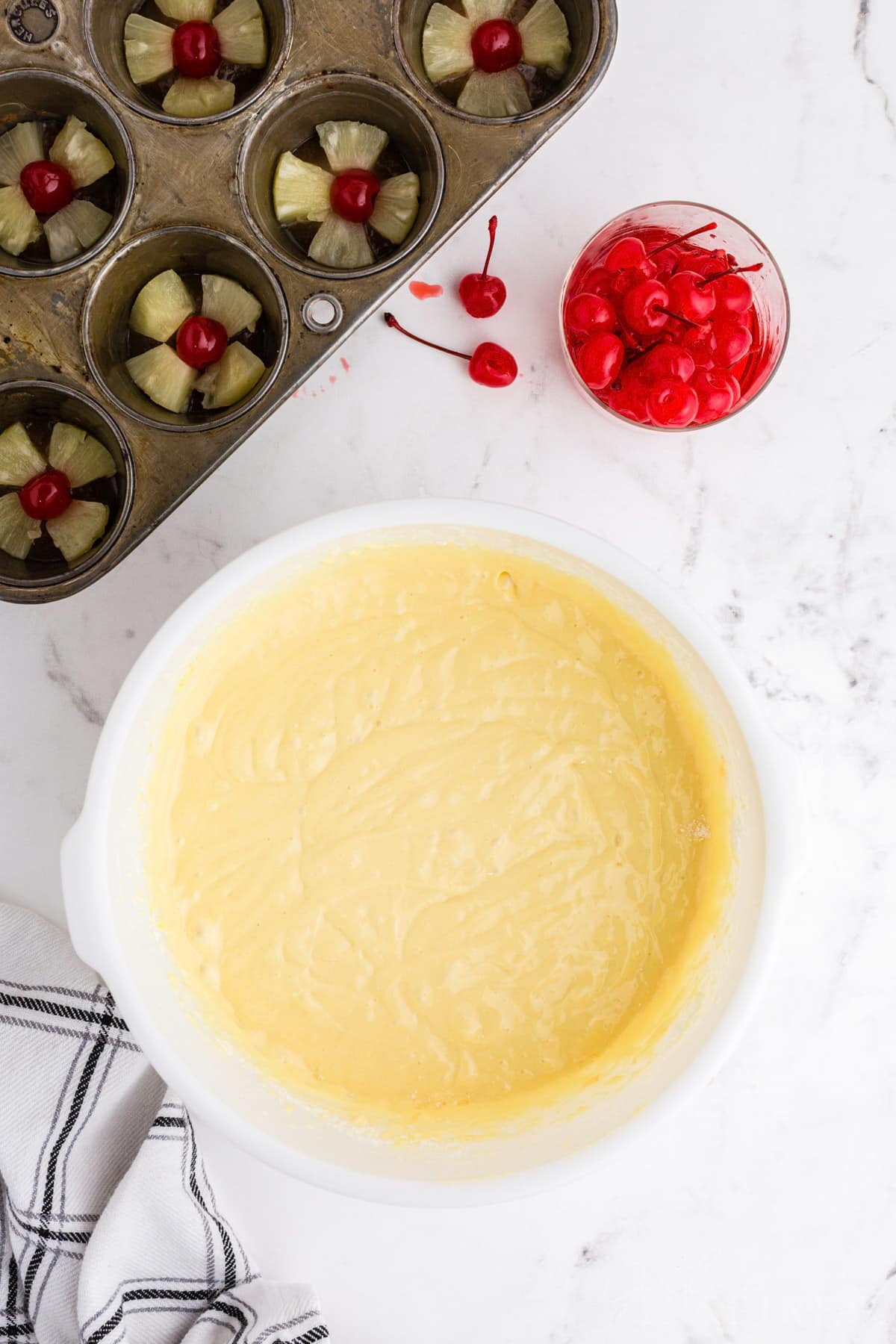 Pineapple upside down cake batter in a large mixing bowl, pan with assembled pineapple slices and maraschino cherries, on a marble countertop. 