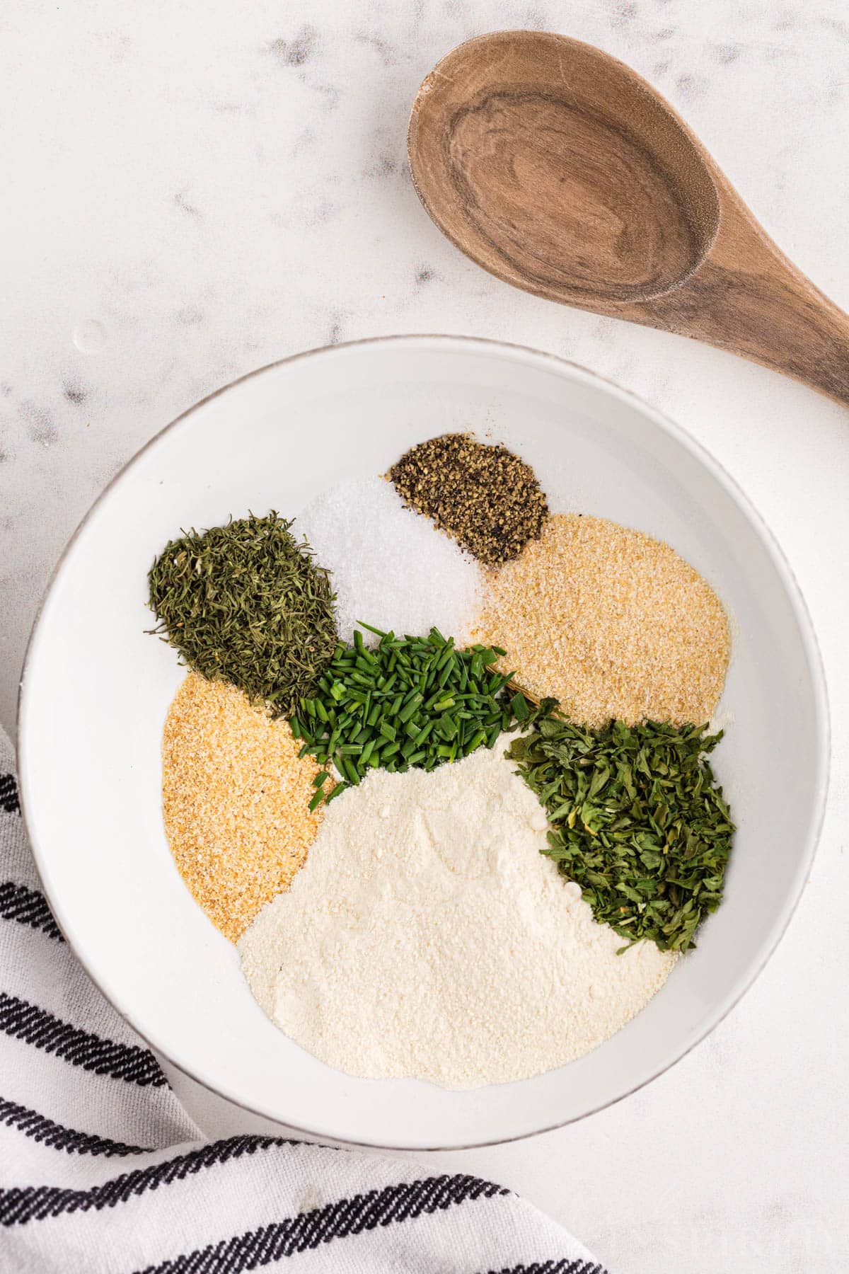 Bowl with ranch seasoning packet ingredients and a wooden spoon on a marble countertop.