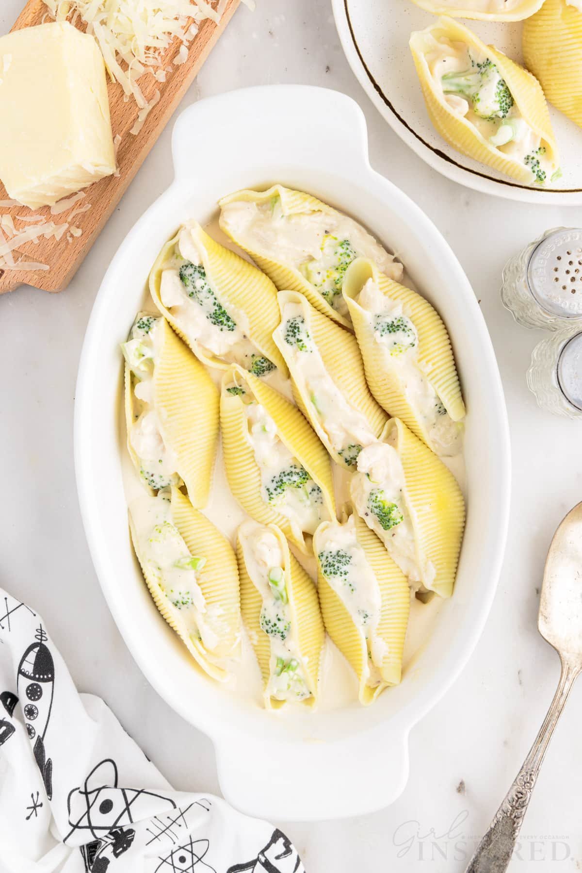 Overhead view of an oval casserole dish of Chicken Alfredo Stuffed Shells next to a plate of shells, mozzarella cheese, and salt and pepper.