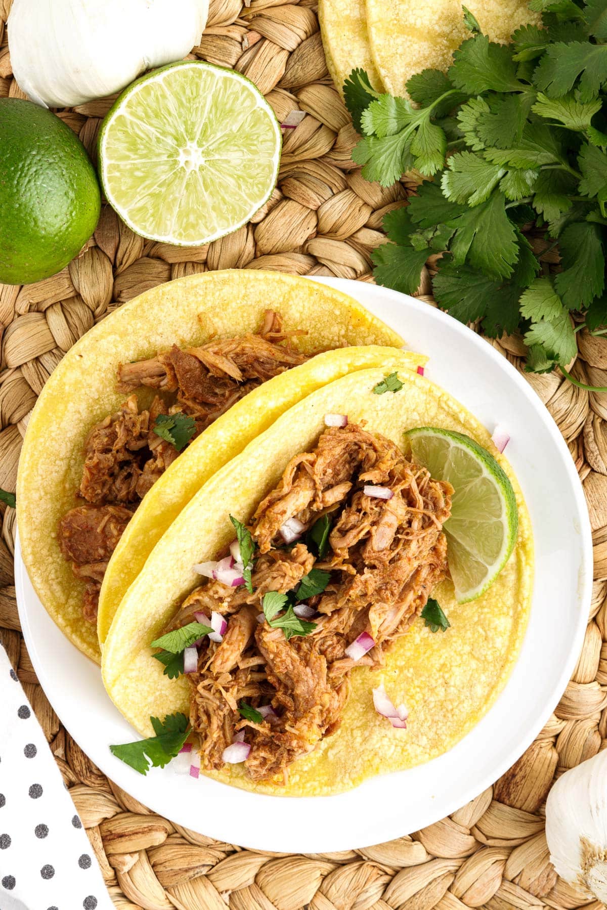 Overhead view of two Pork Barbacoa tacos on a plate next to limes and cilantro.