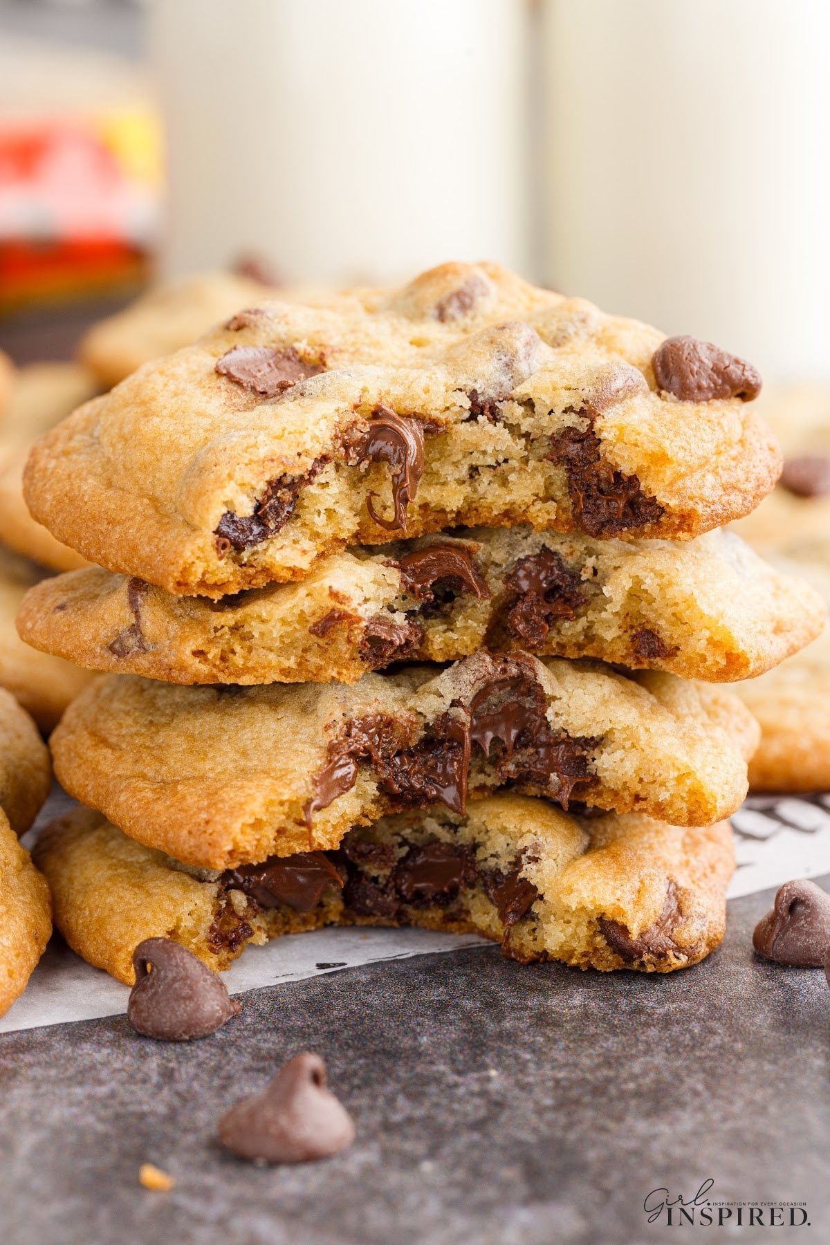 A stack of Toll house chocolate chip cookies with bites taken out and glasses of milk in the background.