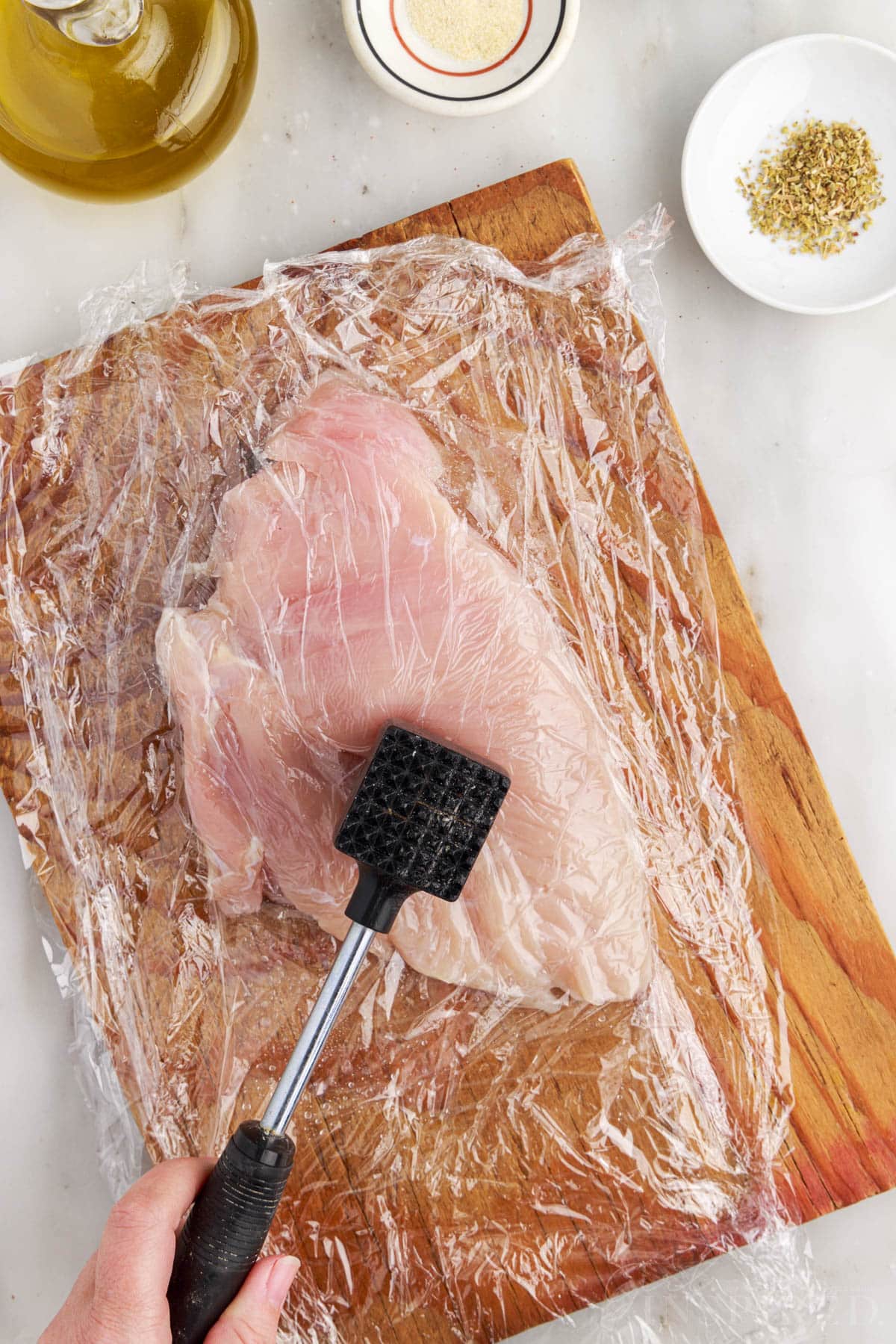 A chicken breast on a cutting board covered with saran wrap pounded with a meat mallet.