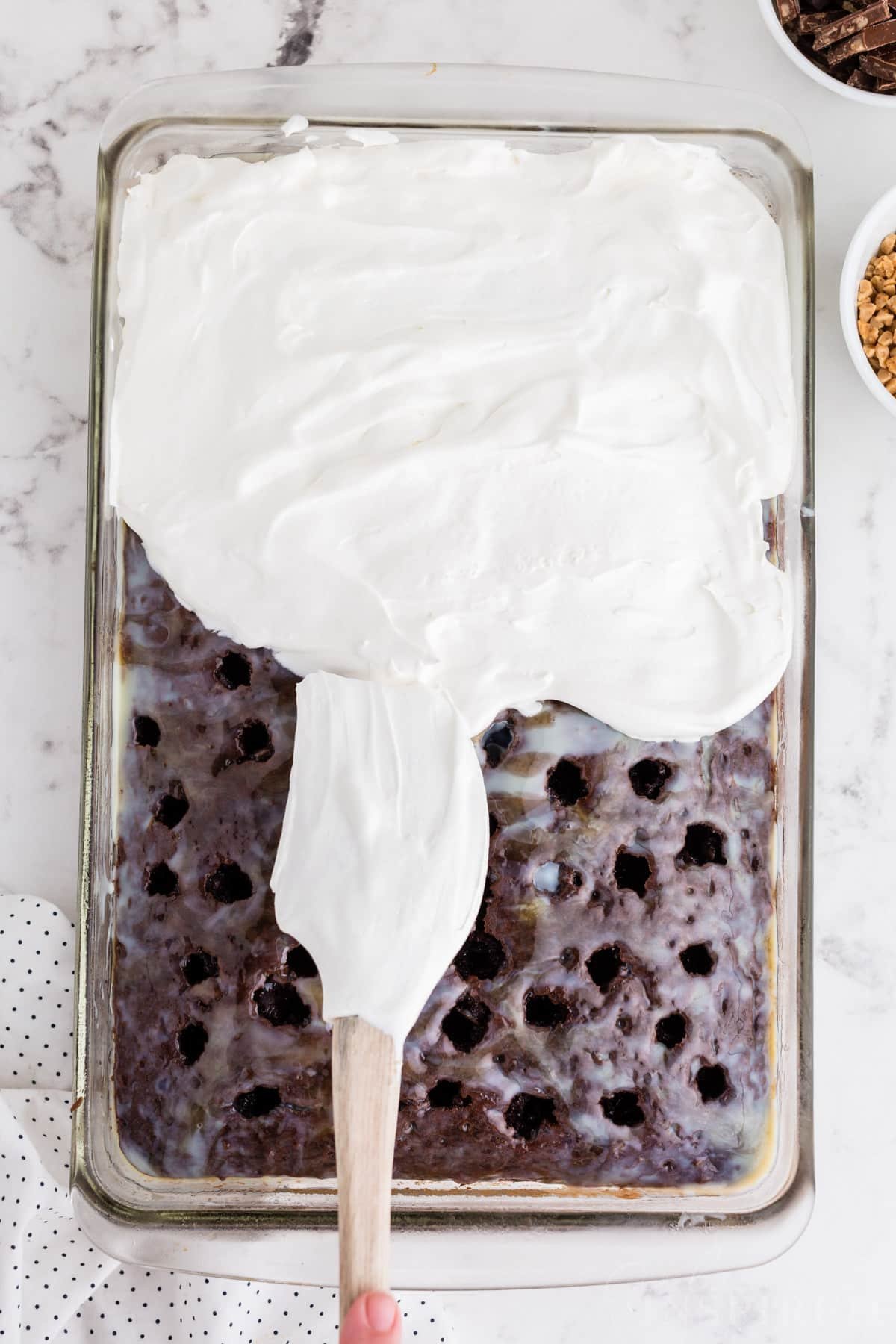 Whipped topping with plastic spatula spread evenly over the poked cake in a glass baking dish on the countertop.