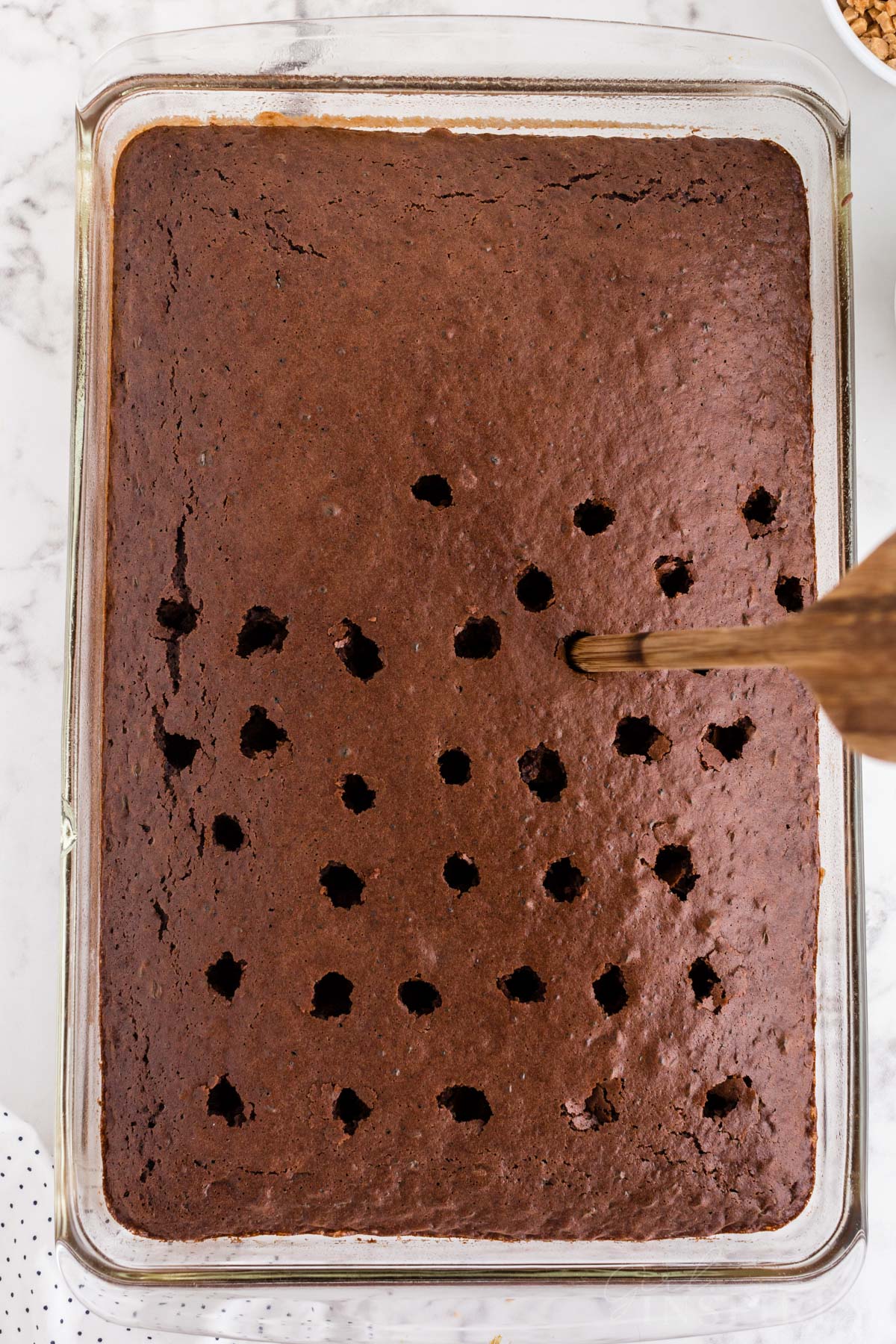 Back of a wooden spoon poking holes in the baked chocolate cake base in a glass baking dish on a countertop.