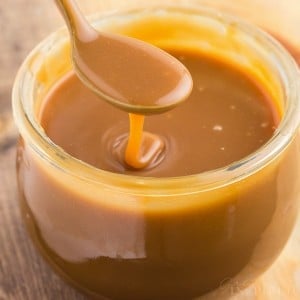 Close up of brown sugar caramel sauce in a glass jar with a metal teaspoon dripping caramel sauce.