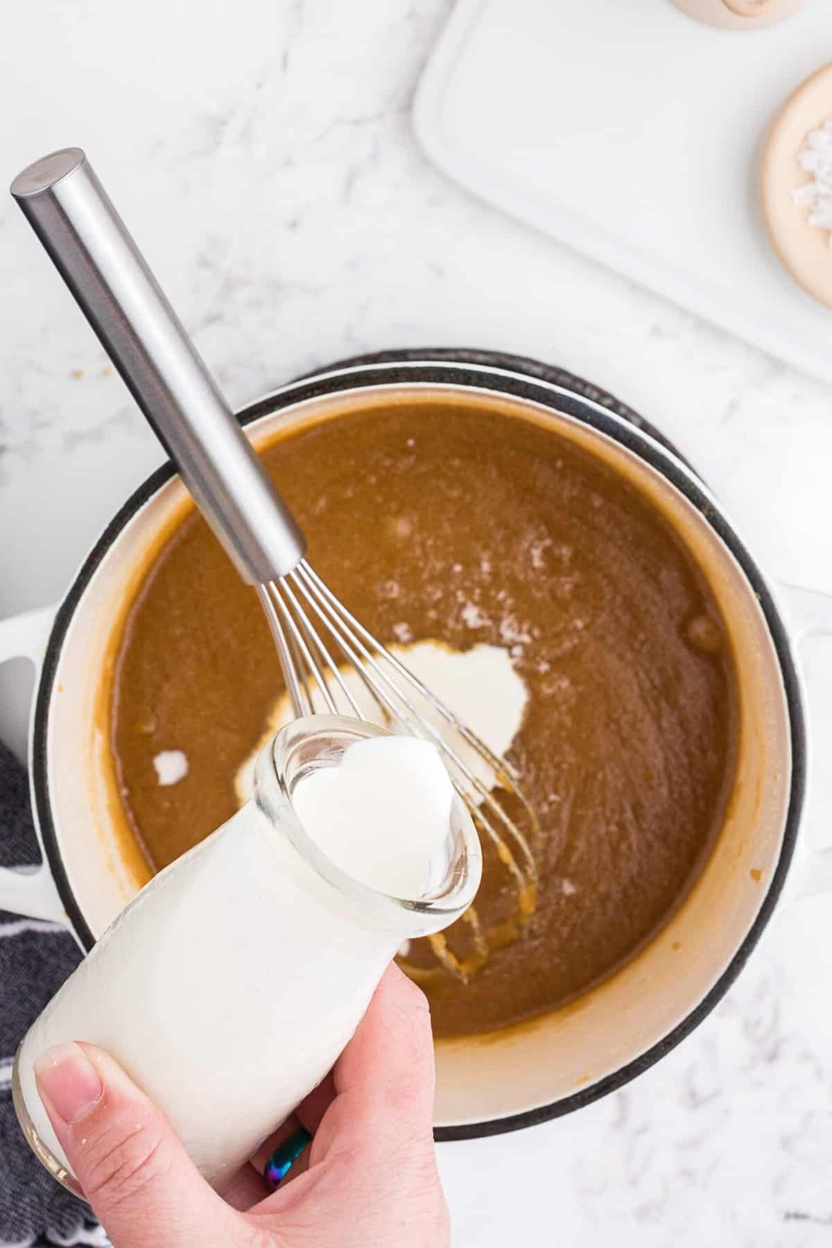 Saucepan with brown sugar and butter mixture, heavy cream poured into the saucepan, metal whisk resting in the pan.