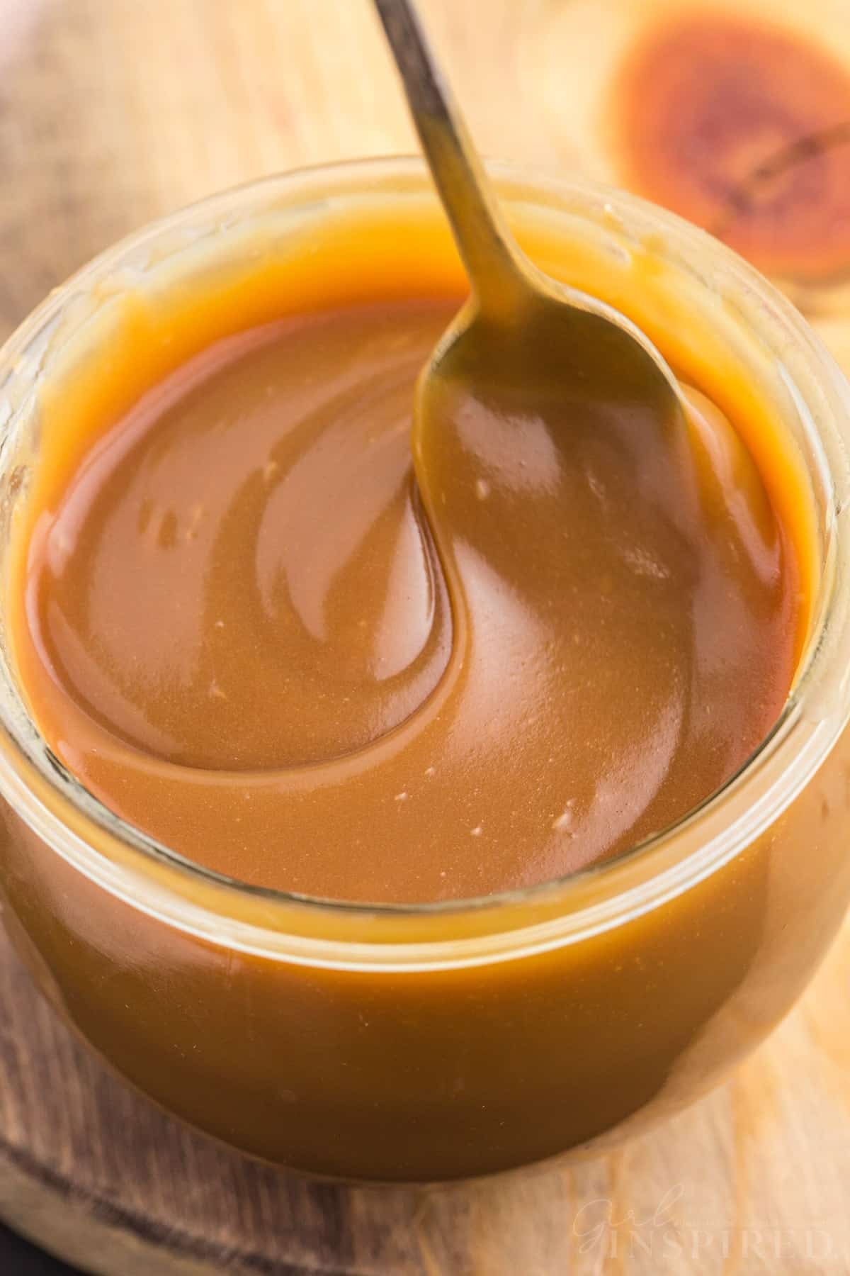 Close up of brown sugar caramel sauce in a glass jar with metal spoon on a wooden kitchen board.