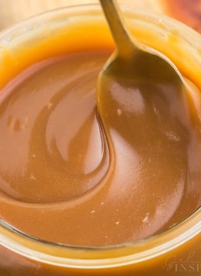 Close up of brown sugar caramel sauce in a glass jar with metal spoon on a wooden kitchen board.